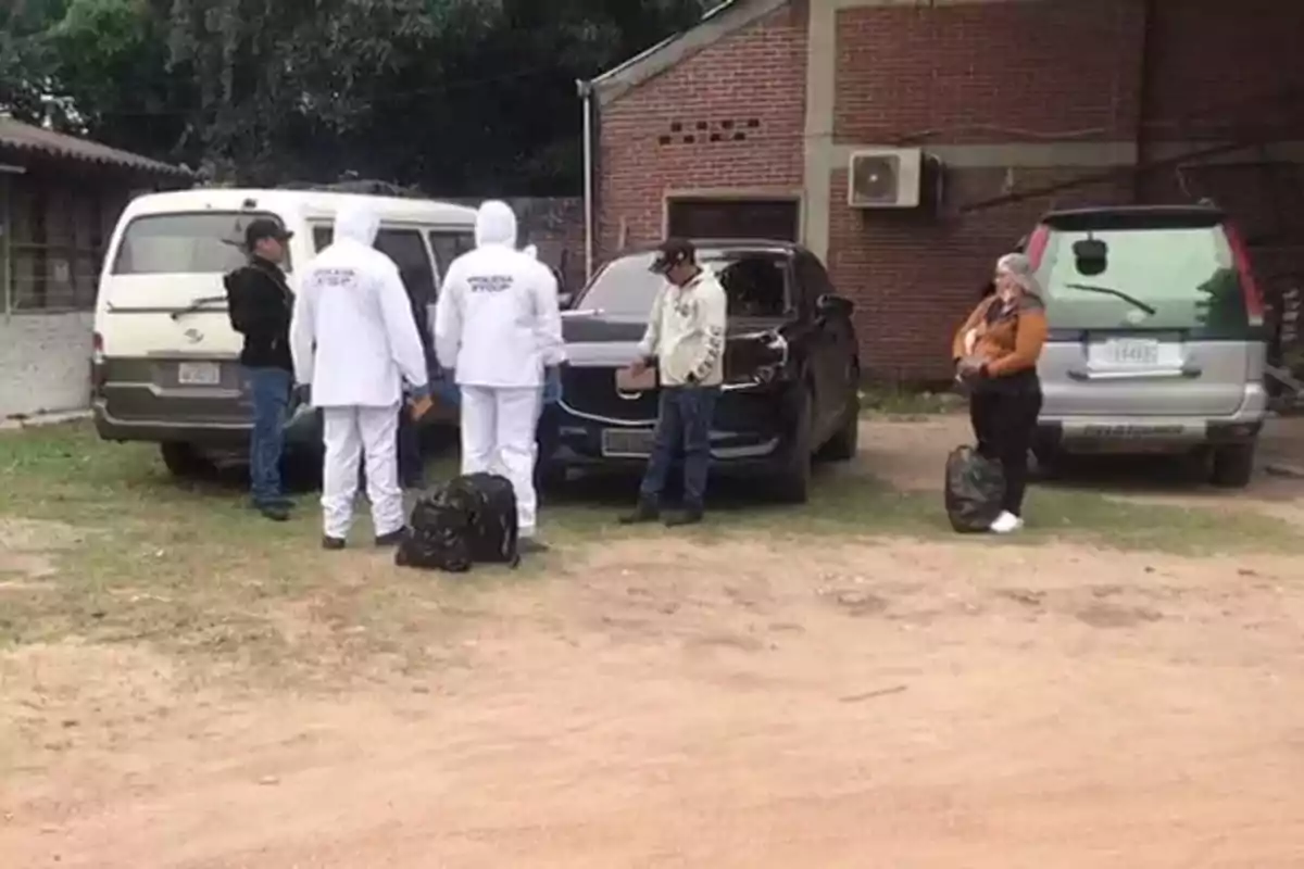 Varias personas, incluyendo dos con trajes blancos de protección, están reunidas junto a tres vehículos estacionados frente a un edificio de ladrillo