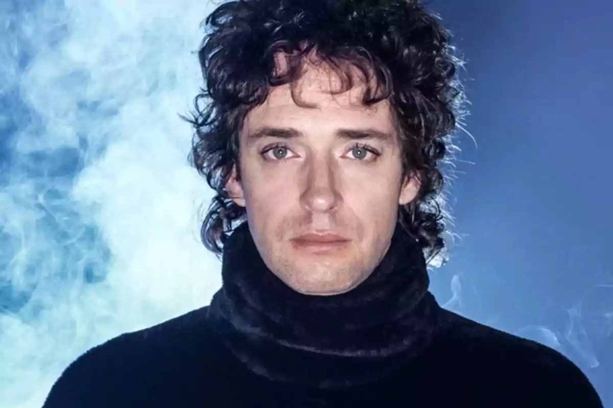 Persona de cabello rizado y oscuro con suéter negro posando frente a un fondo azul con humo Persona de cabello rizado y oscuro con suéter negro posando frente a un fondo azul con humo