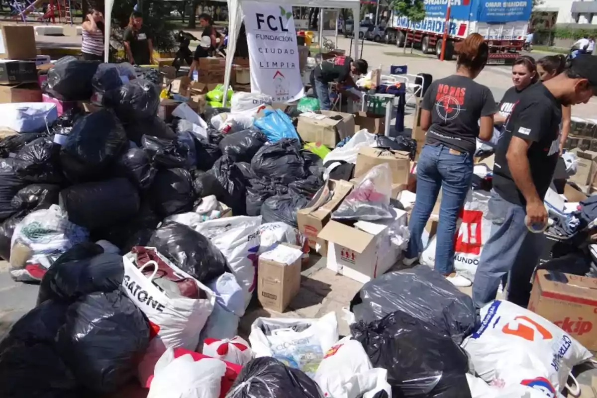 Personas organizan y clasifican donaciones de ropa y artículos en un espacio al aire libre, rodeados de bolsas y cajas.