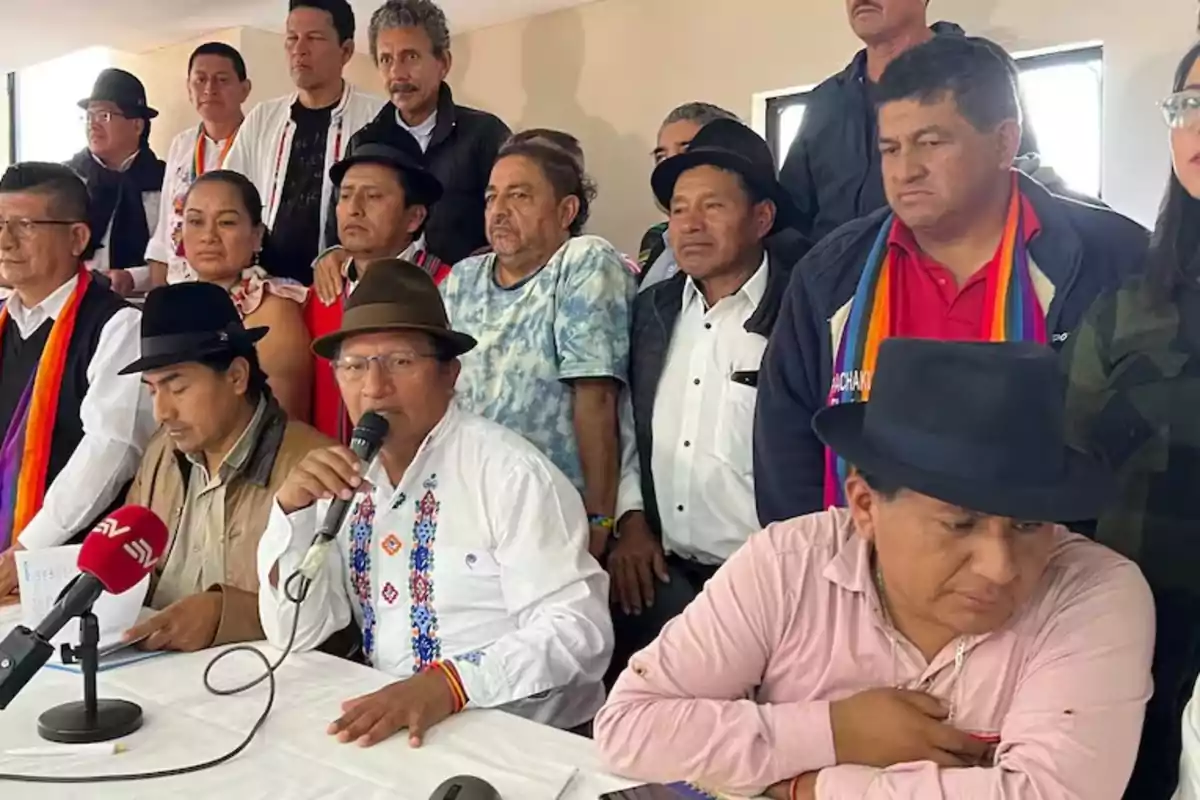 A group of people, some with hats, is gathered at a press conference; one person is speaking into a microphone while others watch.