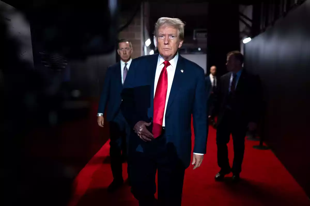 Blond-haired man in a blue suit and red tie walking on a red carpet accompanied by other people in a dark hallway Blond-haired man in a blue suit and red tie walking on a red carpet accompanied by other people in a dark hallway