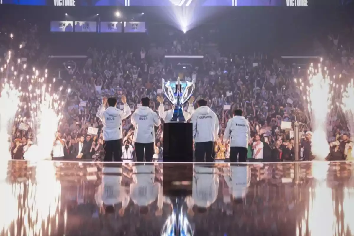 Un equipo de esports celebra en el escenario con un trofeo grande mientras el público aplaude y hay fuegos artificiales a su alrededor.