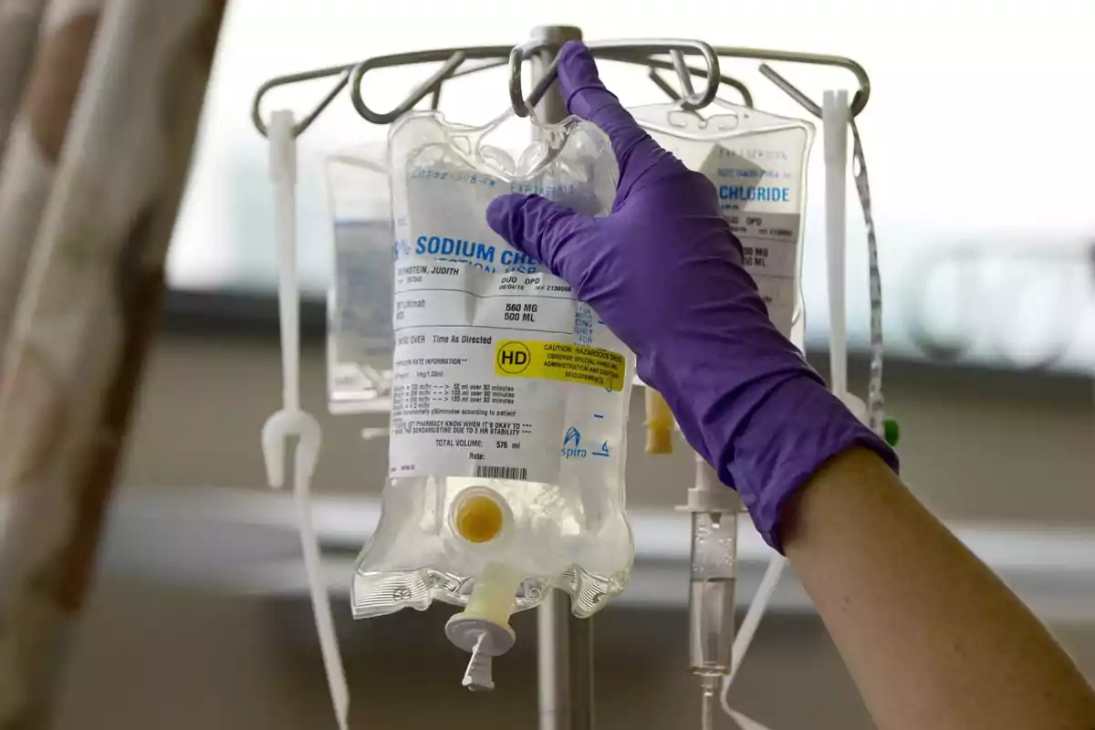 Una mano con guante morado ajusta una bolsa de solución salina en un soporte de suero en un entorno médico. Una mano con guante morado ajusta una bolsa de solución salina en un soporte de suero en un entorno médico.