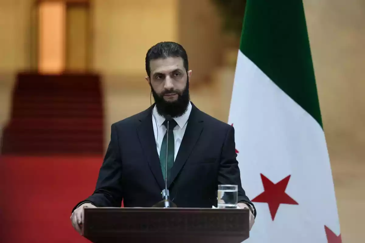 Un hombre de traje oscuro y corbata verde está de pie detrás de un podio con un micrófono, junto a una bandera con estrellas rojas y un triángulo verde.