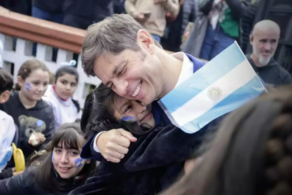 Un hombre sonriente abraza a una niña mientras sostiene una bandera de Argentina rodeado de niños.