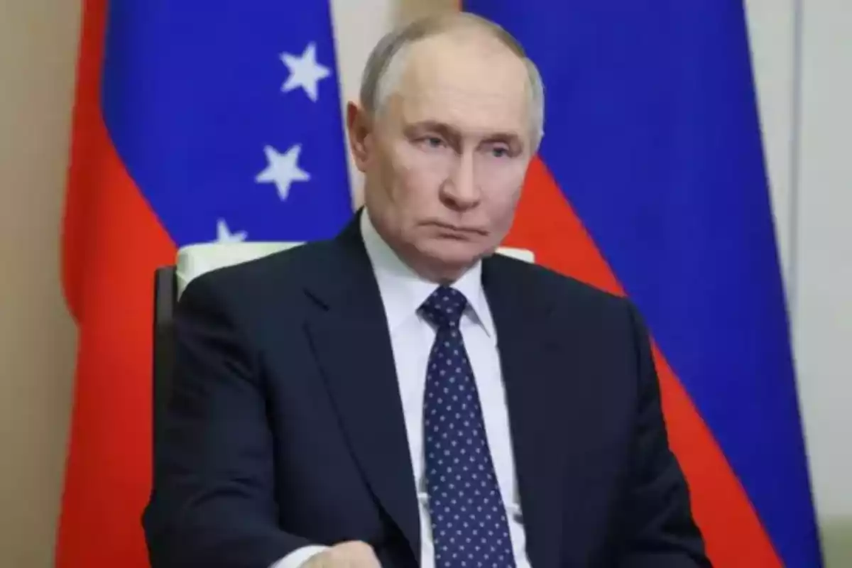 A man in a dark suit and a blue tie with white dots is sitting in front of flags in the background.