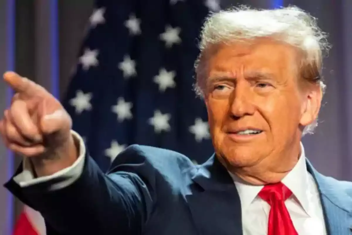 A man in a dark suit and red tie points with his finger while standing in front of a flag with stars.