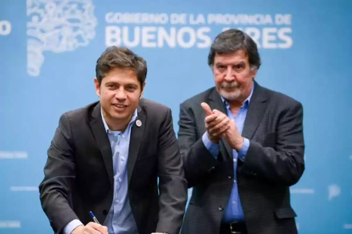 Dos hombres en un evento oficial con un fondo azul que dice "Gobierno de la Provincia de Buenos Aires" uno de ellos está firmando un documento mientras el otro aplaude.