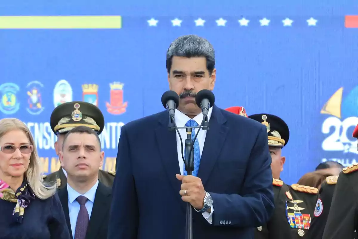 Un hombre en traje oscuro habla frente a varios micrófonos rodeado de personas con uniformes militares y una mujer con gafas en un evento oficial con un fondo azul.