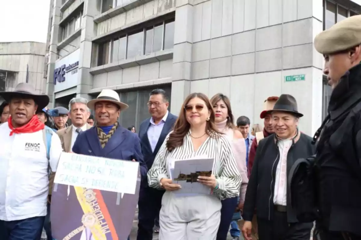 Un grupo de personas camina por la calle frente a un edificio, algunas llevan carteles y documentos.