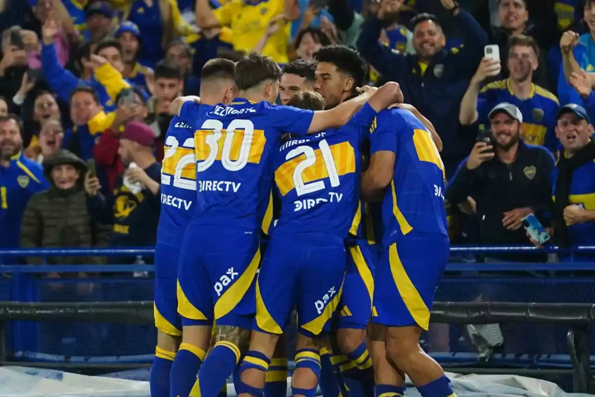 Jugadores de fútbol con uniformes azules y amarillos celebran en grupo mientras los aficionados en el fondo aplauden y toman fotos.