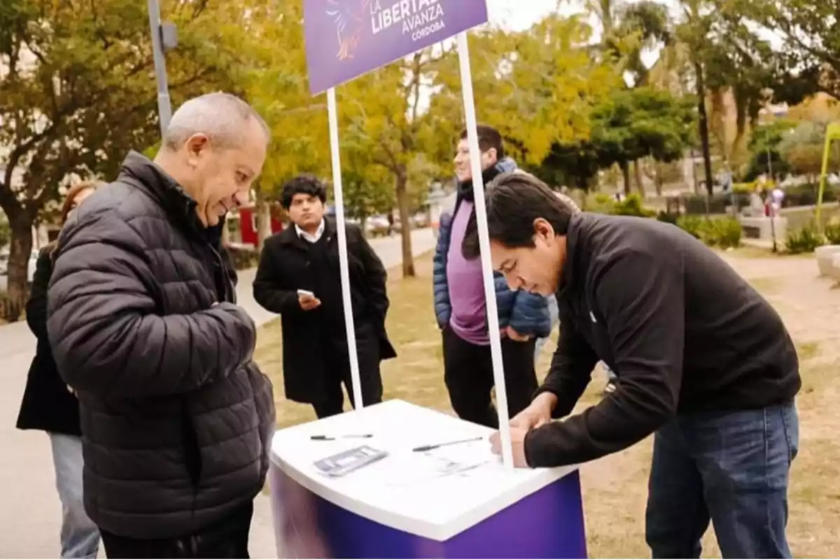 Un hombre firma un papel en una mesa de un stand político al aire libre mientras otras personas observan en un parque Un hombre firma un papel en una mesa de un stand político al aire libre mientras otras personas observan en un parque