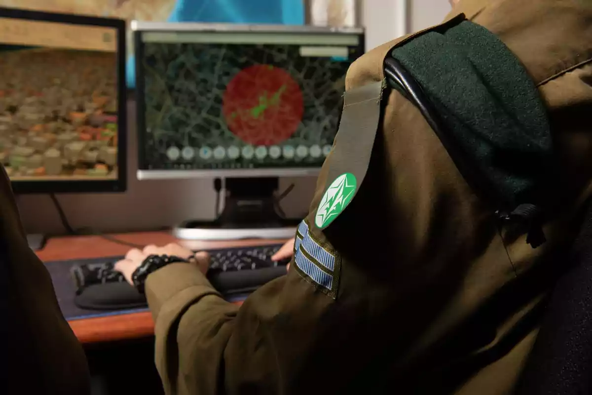 Un soldado en uniforme militar trabaja en una computadora con dos monitores que muestran mapas detallados.