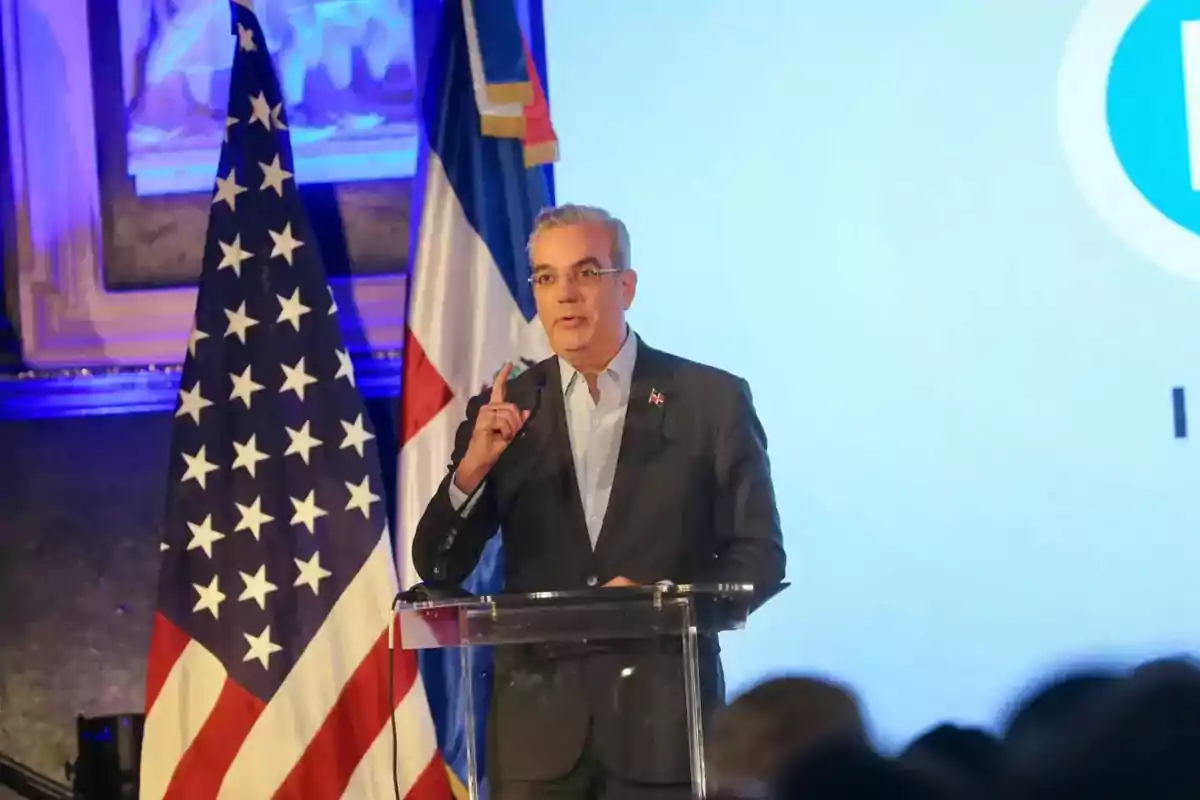 Hombre de traje hablando en un podio con las banderas de Estados Unidos y República Dominicana al fondo