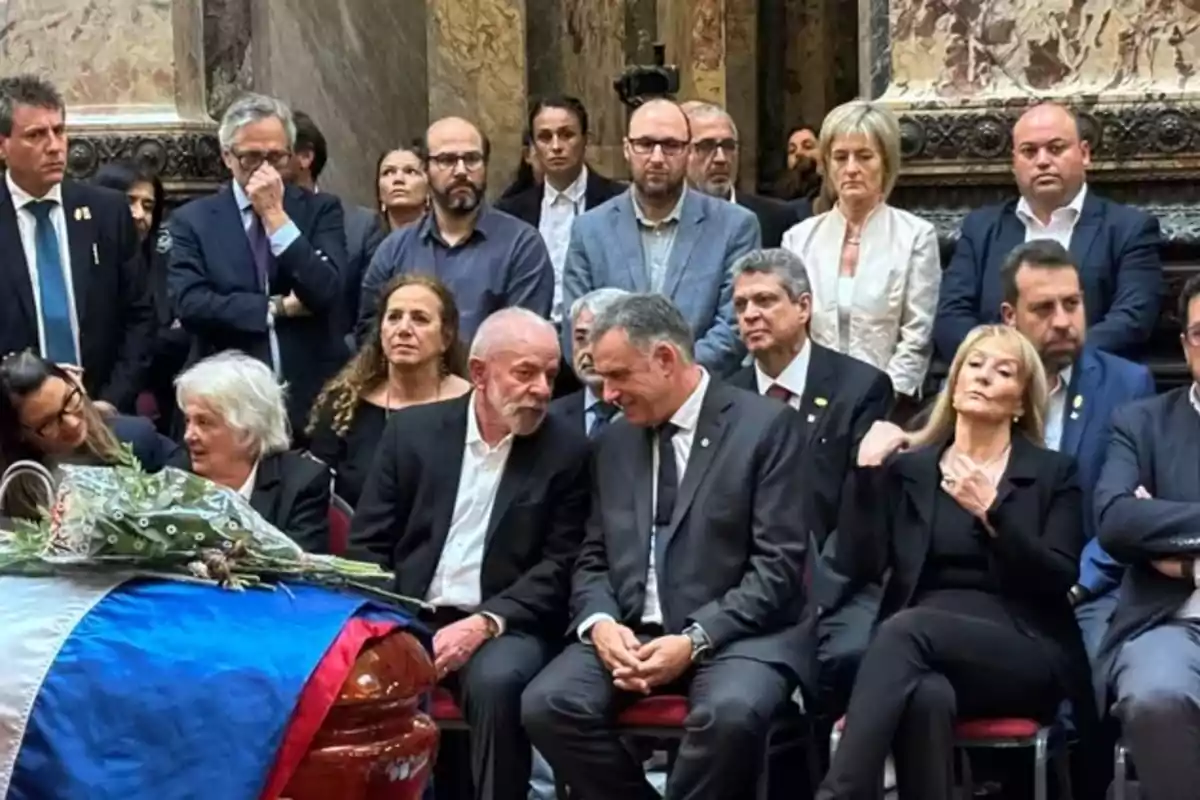 A group of people dressed in black is gathered in a solemn setting around a coffin covered with a flag and flowers. A group of people dressed in black is gathered in a solemn setting around a coffin covered with a flag and flowers.