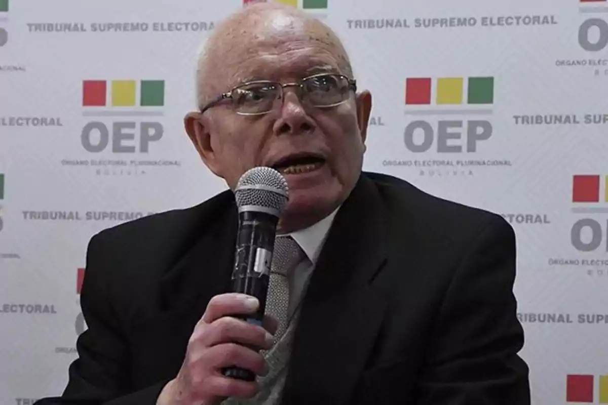 An elderly man with glasses and a dark suit holds a microphone while speaking in front of a background with logos of the Supreme Electoral Court of Bolivia.