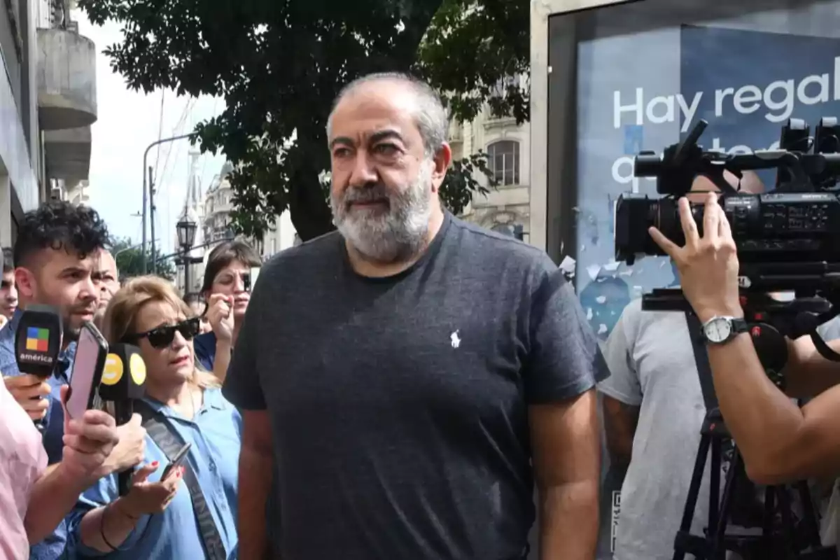 A man with gray hair and a beard wears a gray t-jersey while being surrounded by journalists and cameras on an urban street.