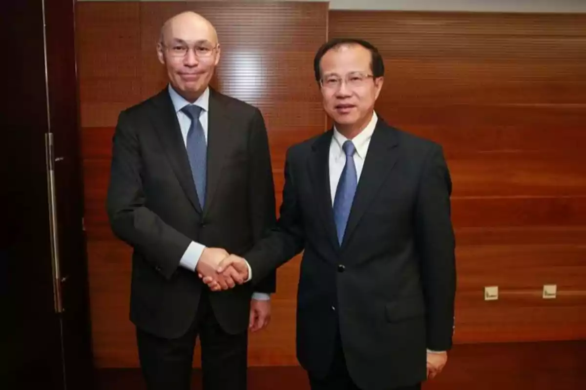 Two men in dark suits shake hands in a formal setting with a wooden background.