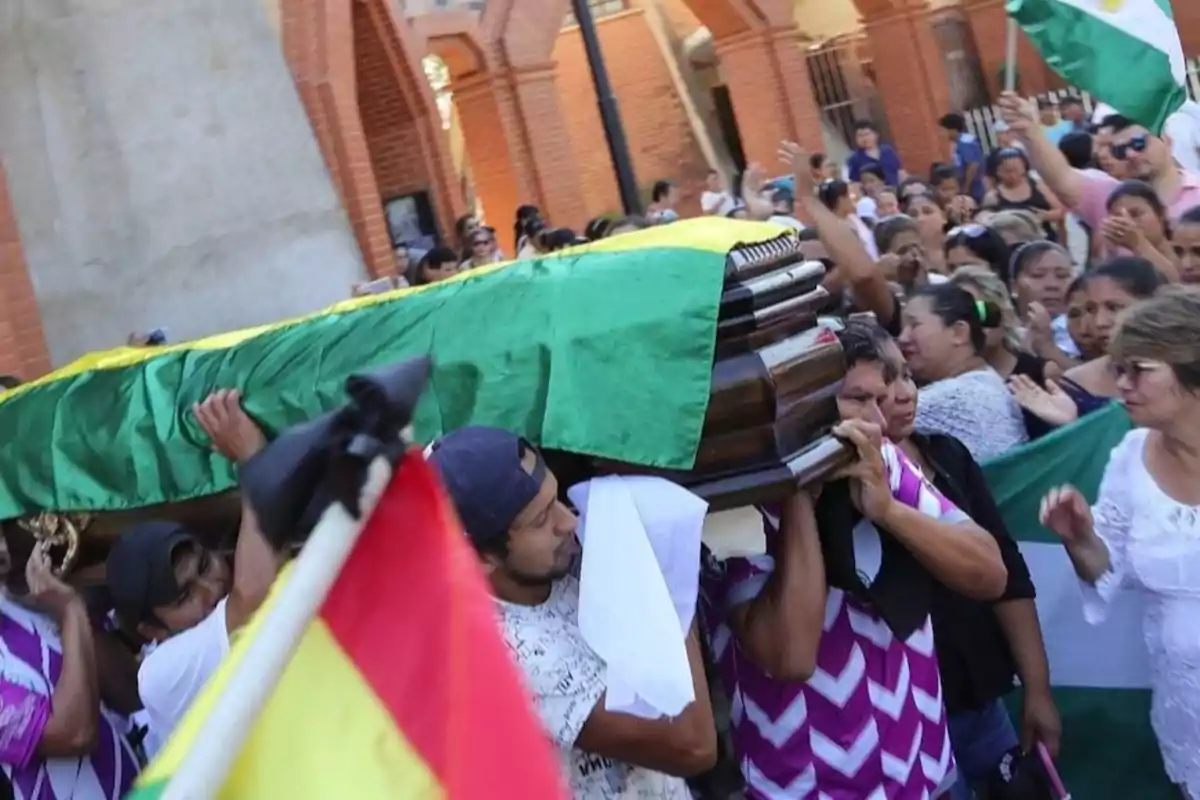 Un grupo de personas lleva un ataúd cubierto con una tela verde y amarilla en una procesión, rodeados de una multitud que sostiene banderas.