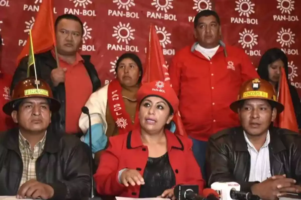 Group of people wearing red clothing and holding red flags, sitting and standing in front of a background with the PAN-BOL logo during a press conference.