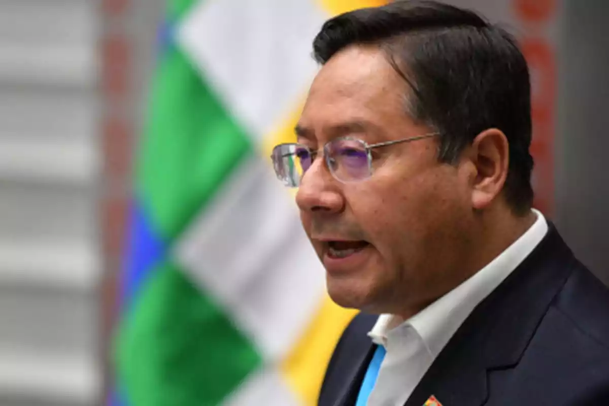 Un hombre con gafas y traje oscuro habla frente a una bandera colorida.