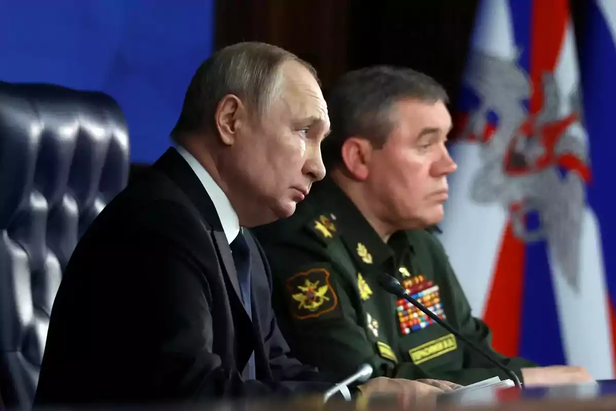 Two men sitting at a table during a conference, one wearing a dark suit and the other in a military uniform, both with serious expressions and flags in the background.