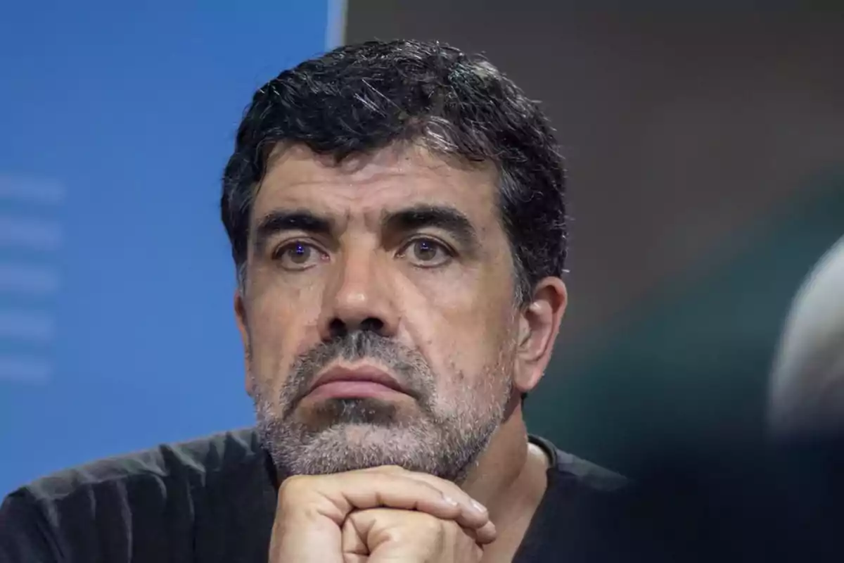 Hombre de cabello oscuro y barba con expresión seria apoyando el mentón sobre su mano Hombre de cabello oscuro y barba con expresión seria apoyando el mentón sobre su mano