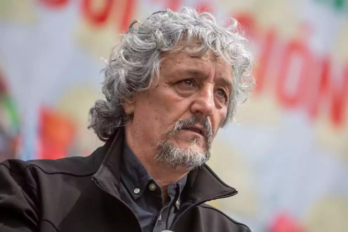 Gray-haired, curly-haired man with a beard and dark jacket looking to the side with a serious expression and a blurred background Gray-haired, curly-haired man with a beard and dark jacket looking to the side with a serious expression and a blurred background