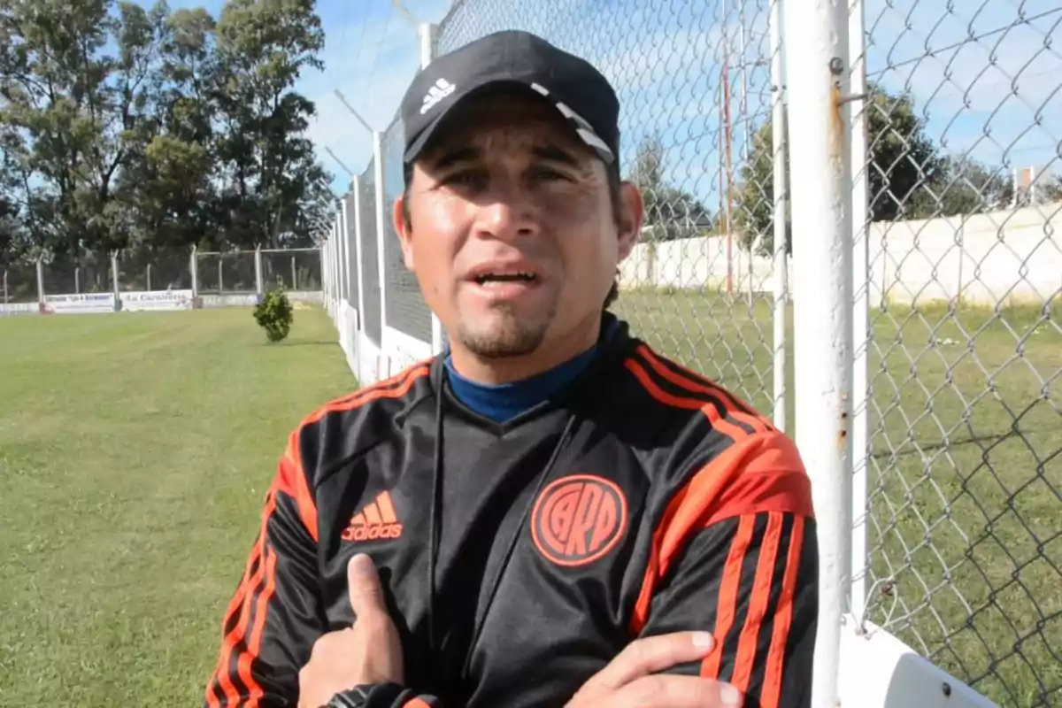 Un hombre con gorra y ropa deportiva de River Plate está de pie junto a una cerca en un campo de fútbol. Un hombre con gorra y ropa deportiva de River Plate está de pie junto a una cerca en un campo de fútbol.