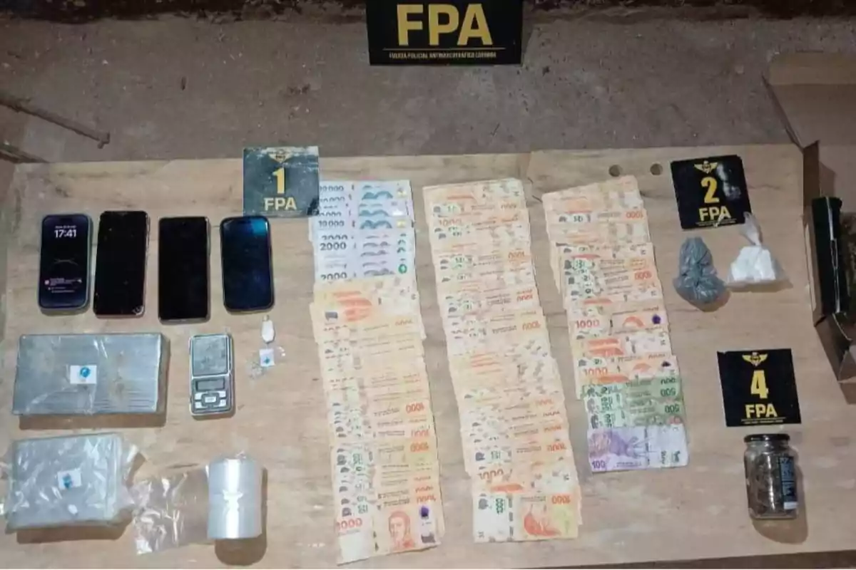 On a wooden table, there are several Argentine bills, four cell phones, a digital scale, packages containing substances, a small jar, plastic bags, and signs from the Córdoba Anti-Narcotics Police Force. On a wooden table, there are several Argentine bills, four cell phones, a digital scale, packages containing substances, a small jar, plastic bags, and signs from the Córdoba Anti-Narcotics Police Force.