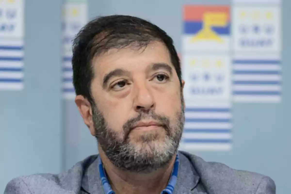 A man with a beard and dark hair, wearing a gray jacket, in front of a background with flags.