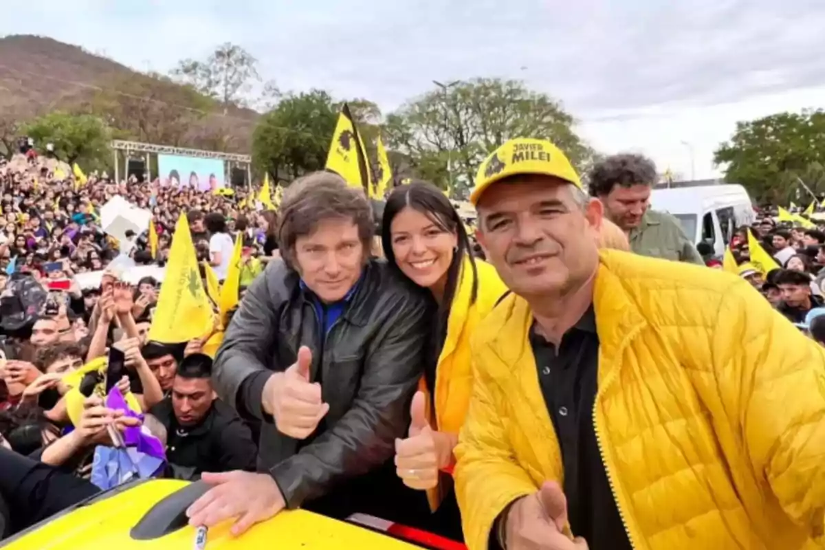 A group of people smiles and gives a thumbs-up gesture at an outdoor event with a crowd and yellow flags in the background.