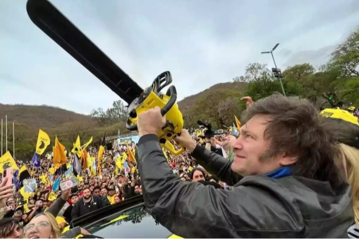 Una persona sostiene una motosierra amarilla en el aire mientras una multitud con banderas lo rodea en un evento al aire libre.