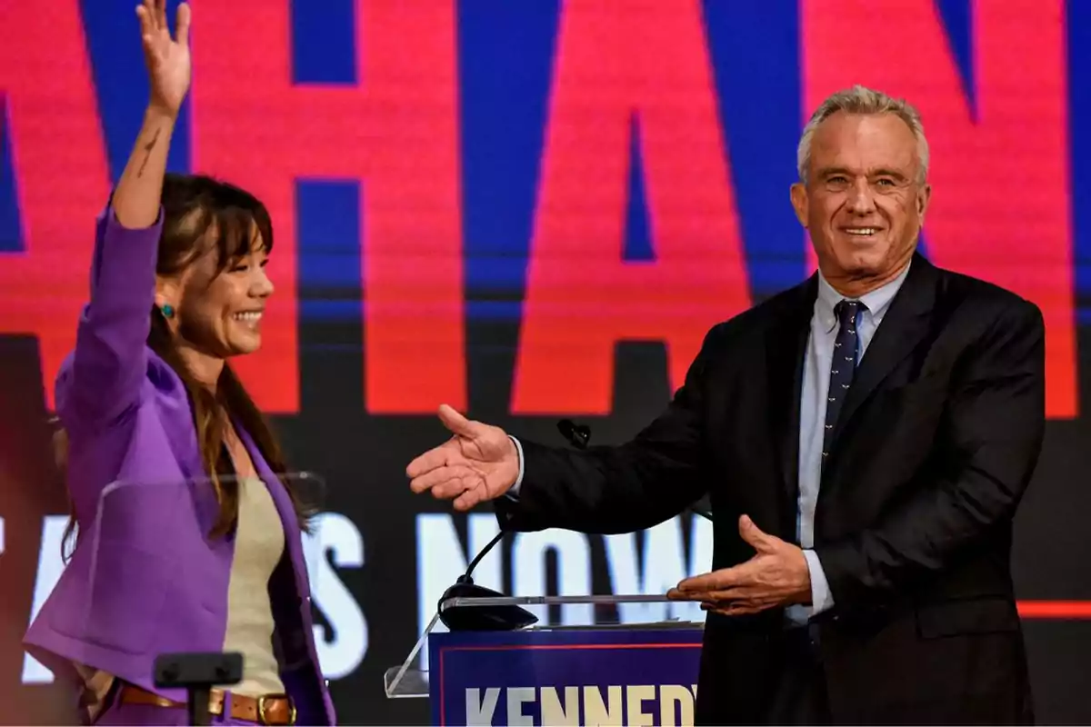 Dos personas en un escenario, una mujer levantando la mano y un hombre sonriendo y extendiendo el brazo hacia ella, con un fondo de letras grandes y coloridas.