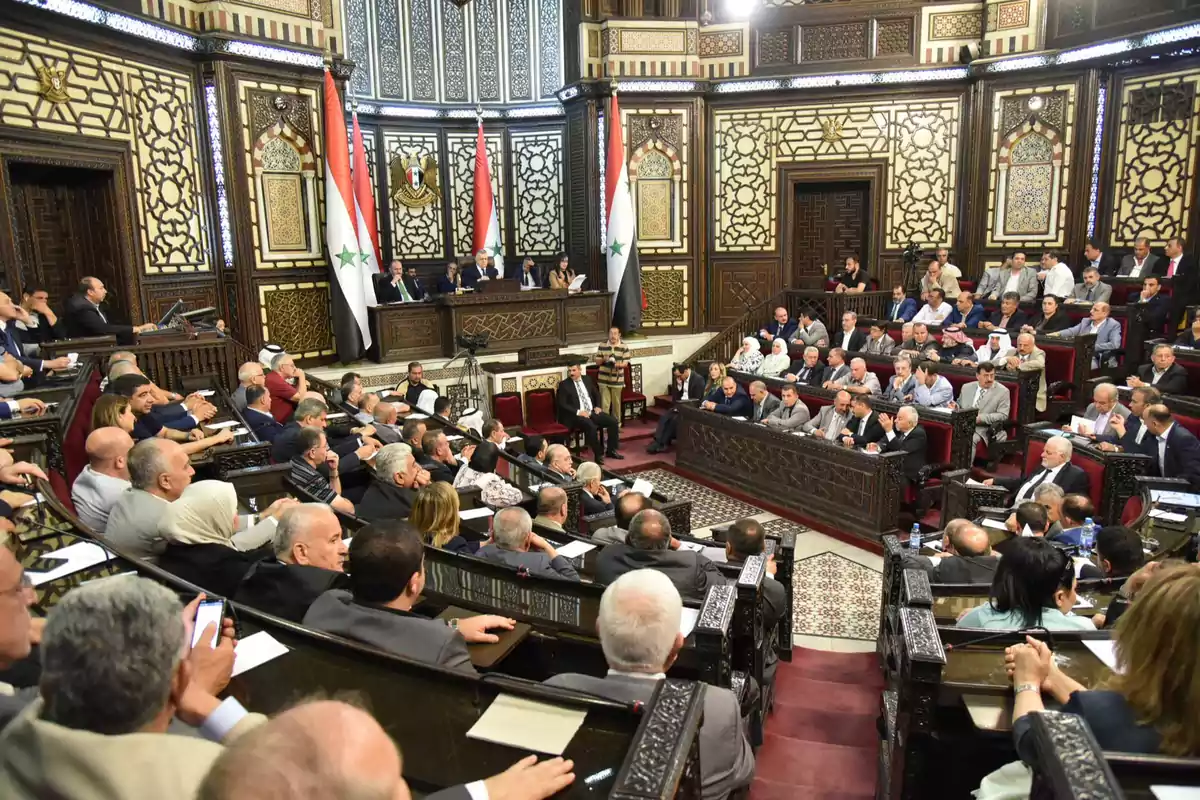 Una asamblea parlamentaria con personas sentadas en un recinto decorado con intrincados diseños geométricos y banderas nacionales. Una asamblea parlamentaria con personas sentadas en un recinto decorado con intrincados diseños geométricos y banderas nacionales.