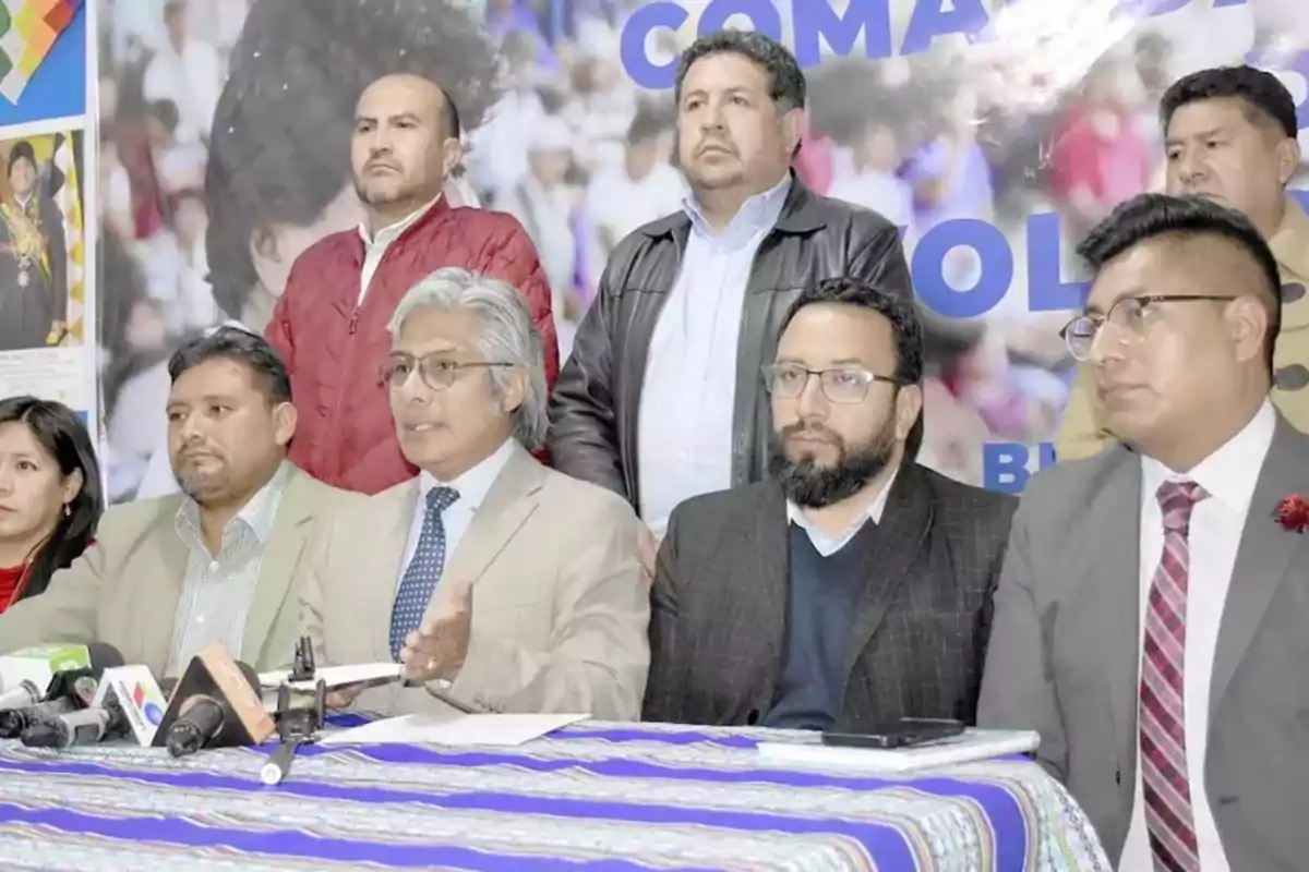 A group of people at a press conference with microphones on a table covered with a colorful tablecloth.