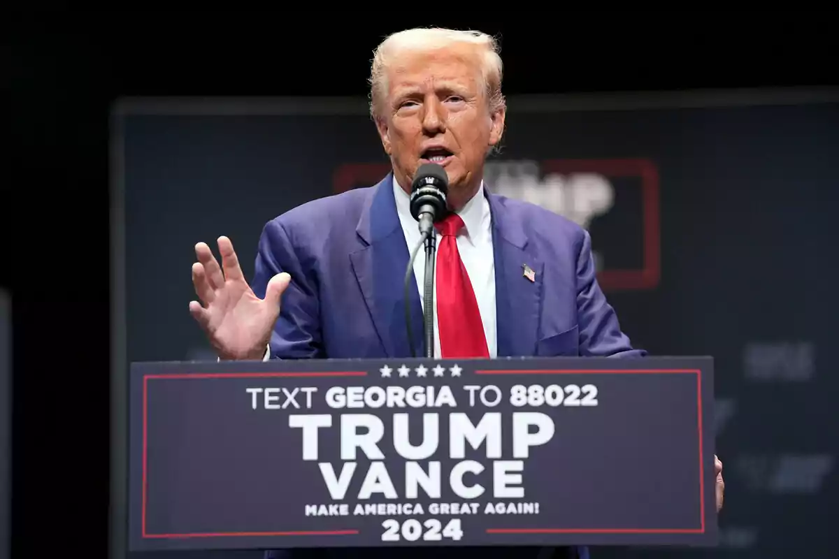 Un hombre hablando en un mitin político con un micrófono y un cartel que dice "TEXT GEORGIA TO 88022 TRUMP VANCE MAKE AMERICA GREAT AGAIN! 2024".