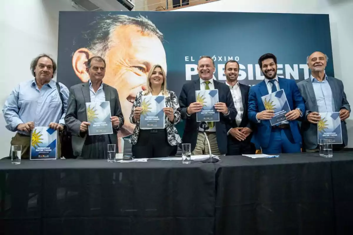 Un grupo de personas posando con folletos en un evento frente a un cartel grande. Un grupo de personas posando con folletos en un evento frente a un cartel grande.