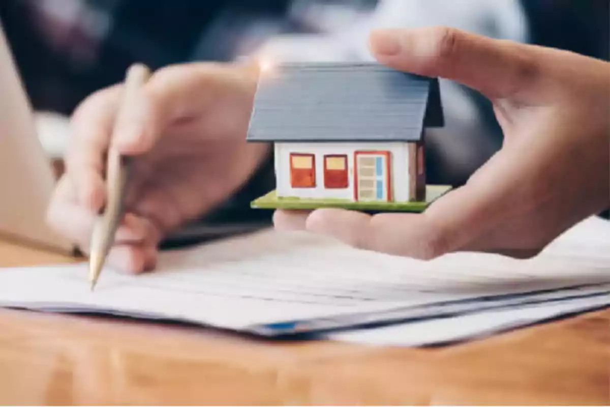 Person holding a model house while signing documents on a table