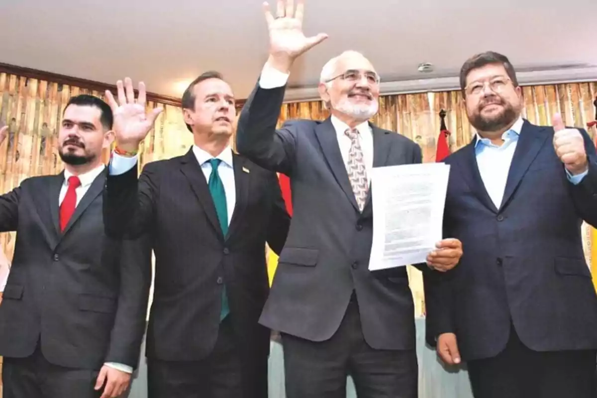 Un grupo de cuatro hombres en trajes formales posan juntos, uno de ellos sostiene un documento mientras levantan las manos en señal de saludo o celebración.