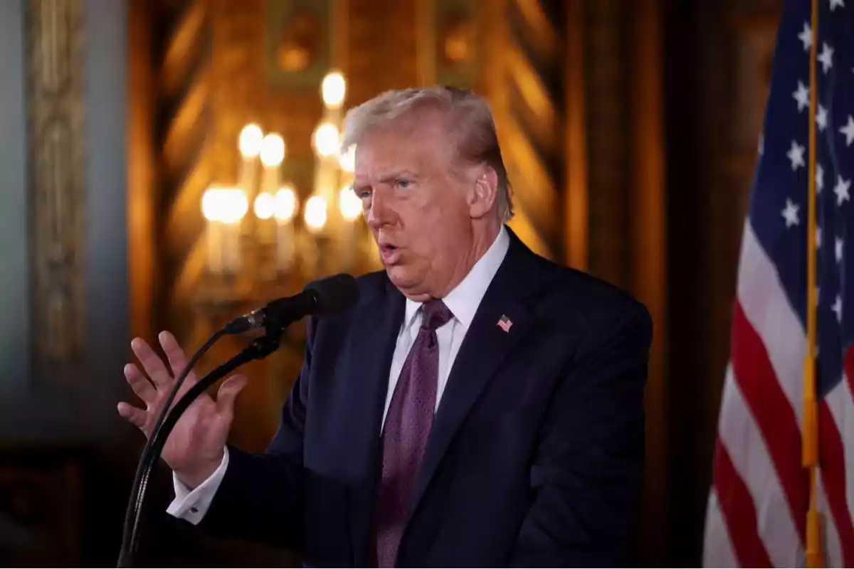 Hombre de cabello rubio con traje oscuro hablando frente a un micrófono junto a una bandera de Estados Unidos en un ambiente elegante con luces de fondo