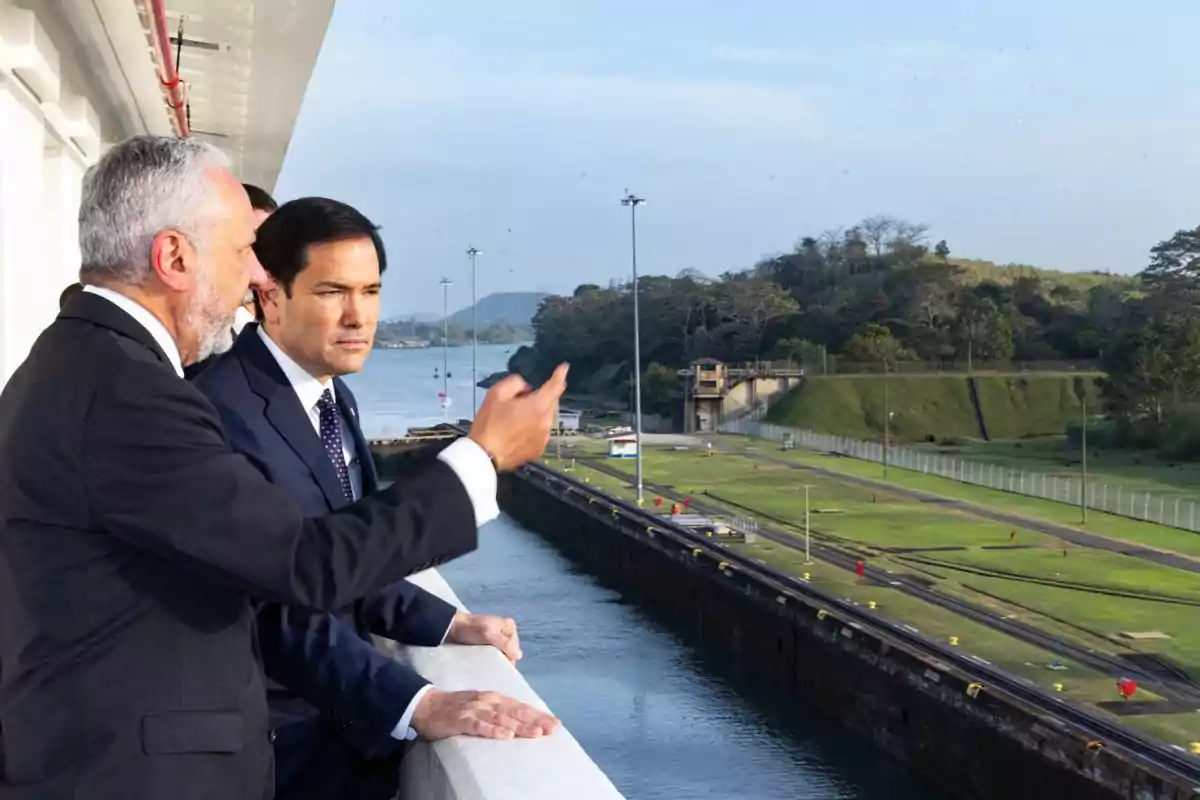 Dos hombres de traje observan el Canal de Panamá desde un balcón.