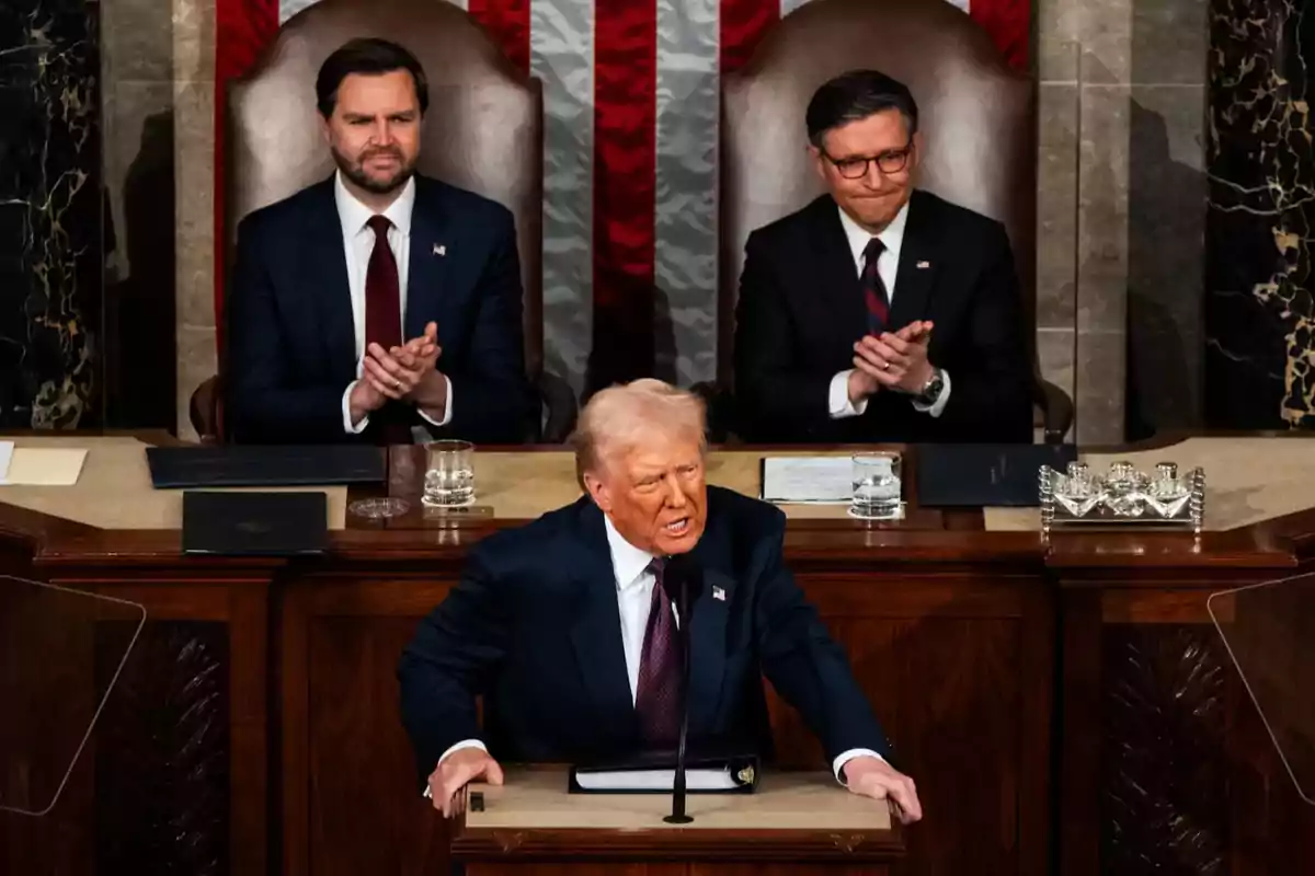 A man speaking at a podium while two people behind him applaud.