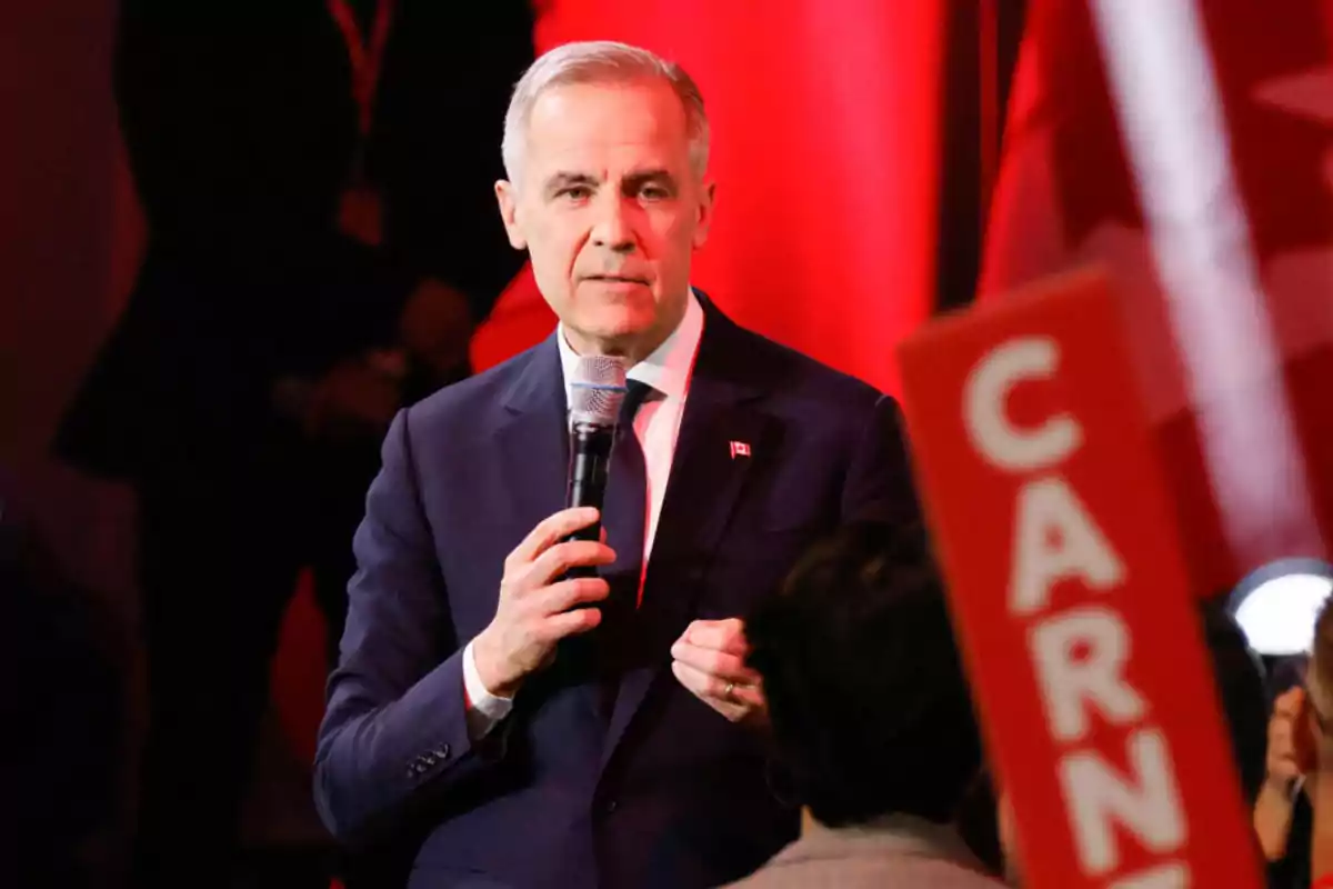 A man in a dark suit holds a microphone while speaking at an event with red lights in the background.