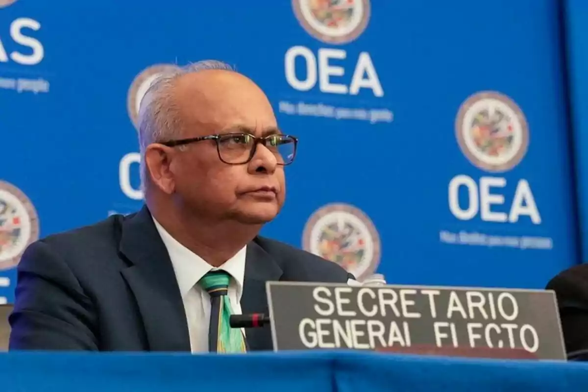 Un hombre con gafas y traje está sentado frente a un micrófono en un evento de la OEA, con un cartel que dice "Secretario General Electo".