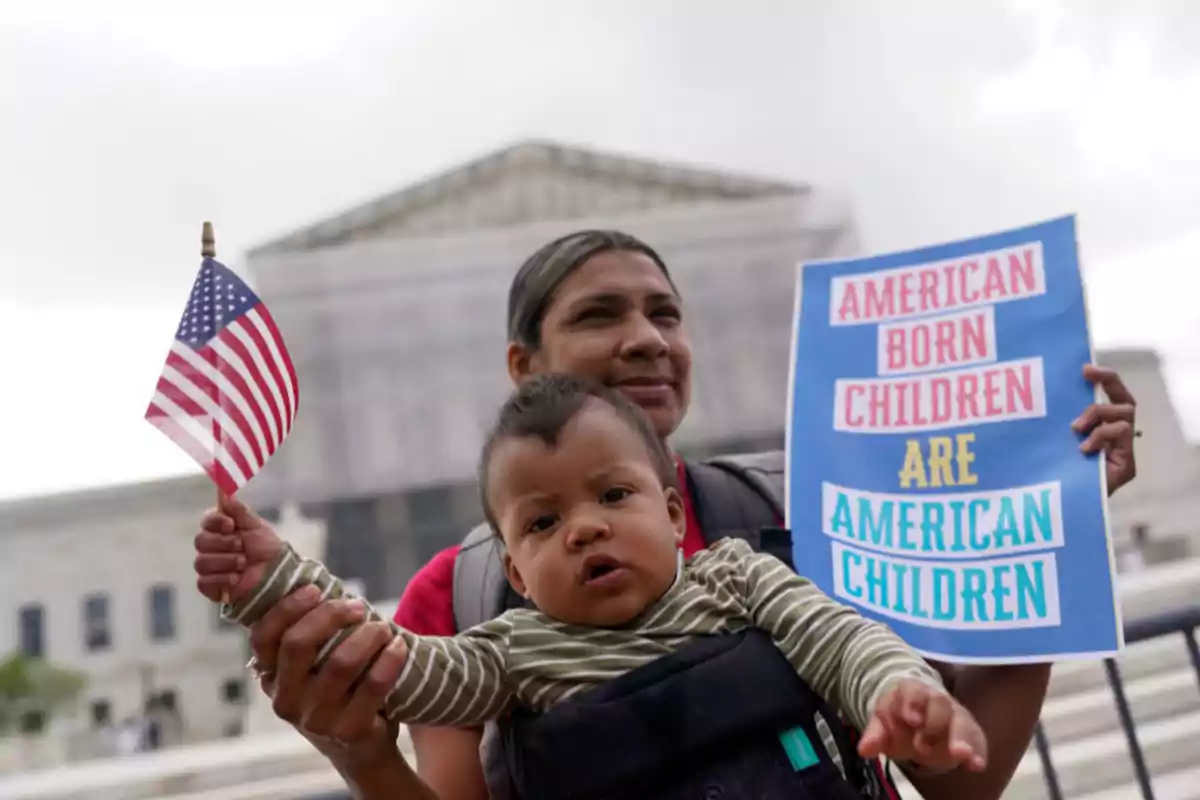 A person holds a baby carrying a United States flag while holding a sign that says A person holds a baby carrying a United States flag while holding a sign that says