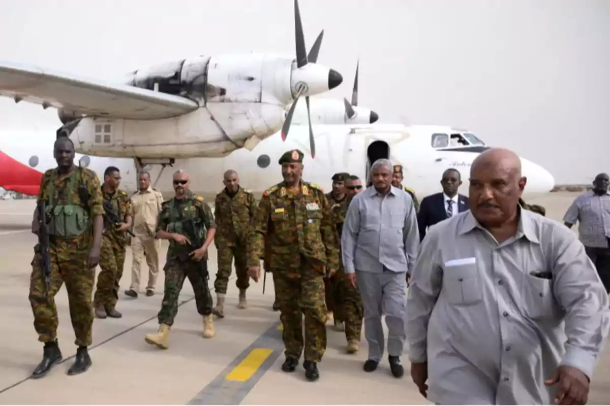 A group of military personnel and civilians walks along the runway of an airport next to a white propeller plane.