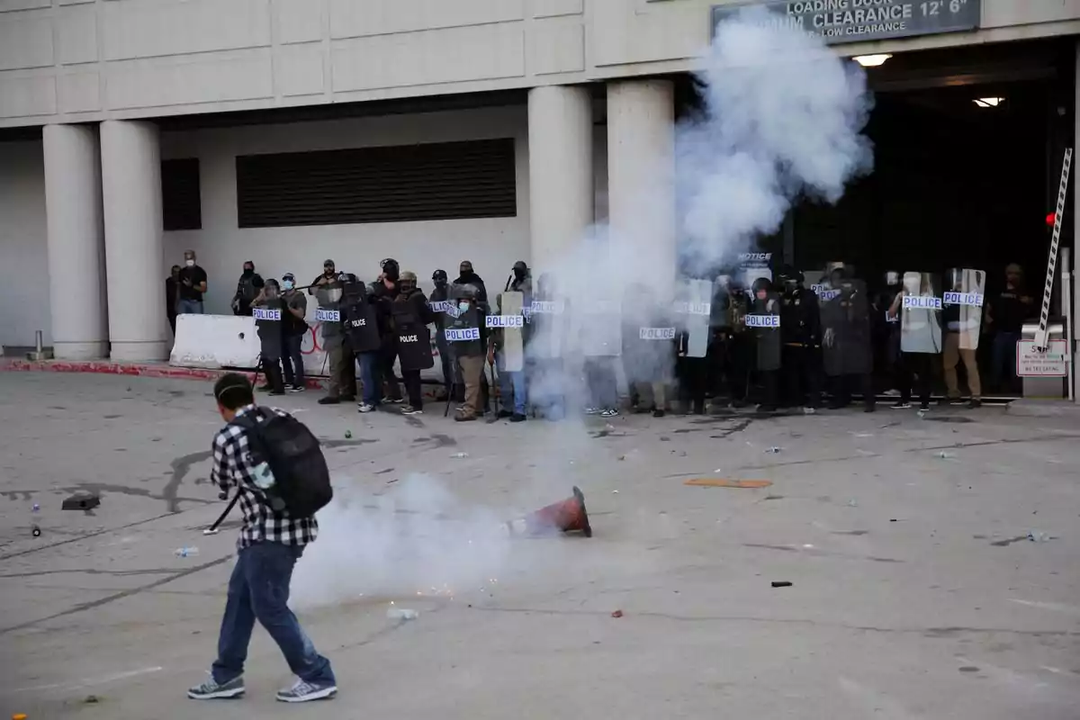 A group of police officers with riot shields is lined up in front of a building while a person with a backpack and a plaid jersey walks near a cloud of smoke on the ground.
