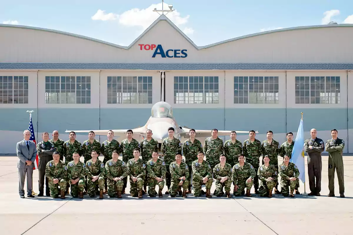 Grupo de militares posando frente a un avión de combate y un hangar con el letrero Top Aces Grupo de militares posando frente a un avión de combate y un hangar con el letrero Top Aces