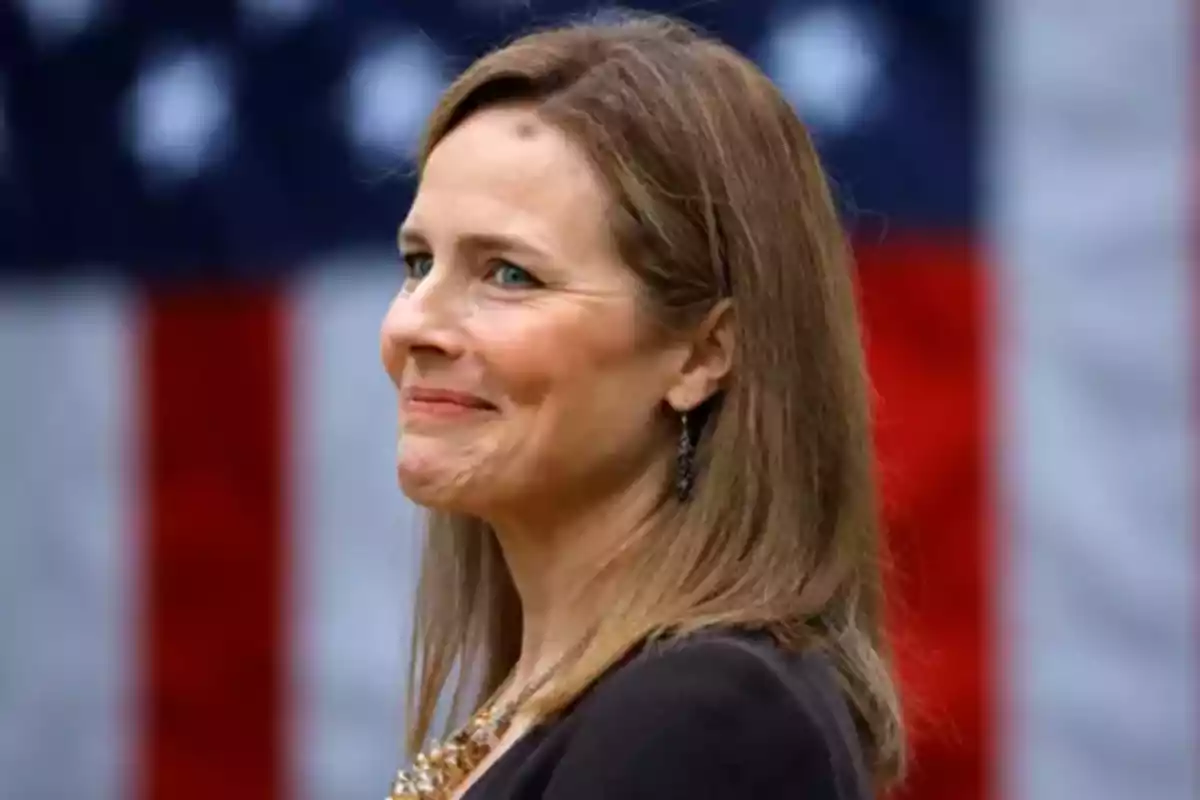 Mujer de cabello castaño claro sonriendo con una bandera de Estados Unidos desenfocada al fondo